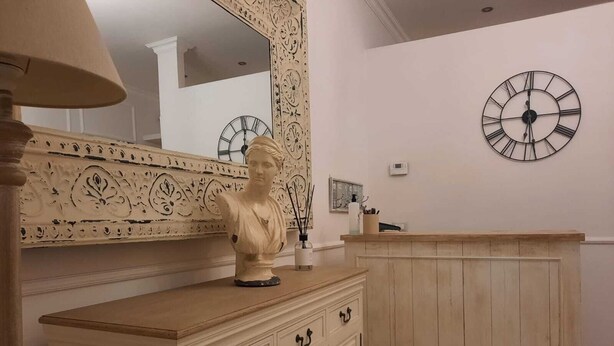 Una habitación con un armario de madera, un busto encima, un espejo grande con detalles ornamentales y un reloj de pared.