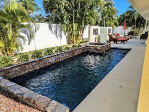 Una piscina con bordes de piedra, rodeada de exuberante vegetación y un muro blanco.