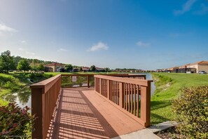 A wooden bridge over a calm water body, surrounded by greenery and residential buildings.