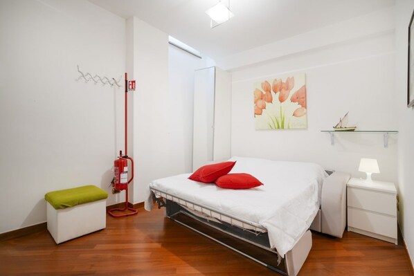 Une chambre à coucher comprenant un lit, une table de chevet, une commode, un banc vert, un extincteur et une étagère fixée au mur.