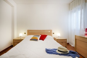 Una cama bien hecha con tablero de madera, dos lámparas de noche, una mesita de noche y un aparador con espejo.