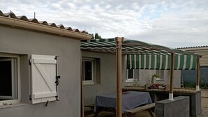 A covered outdoor seating area with a table and chairs, a pergola with a striped awning, and a small garden with a potted plant. - Vias beach.fisherman's house (Vias)