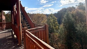 A wooden deck with a staircase leading to a covered area, surrounded by a forested landscape.