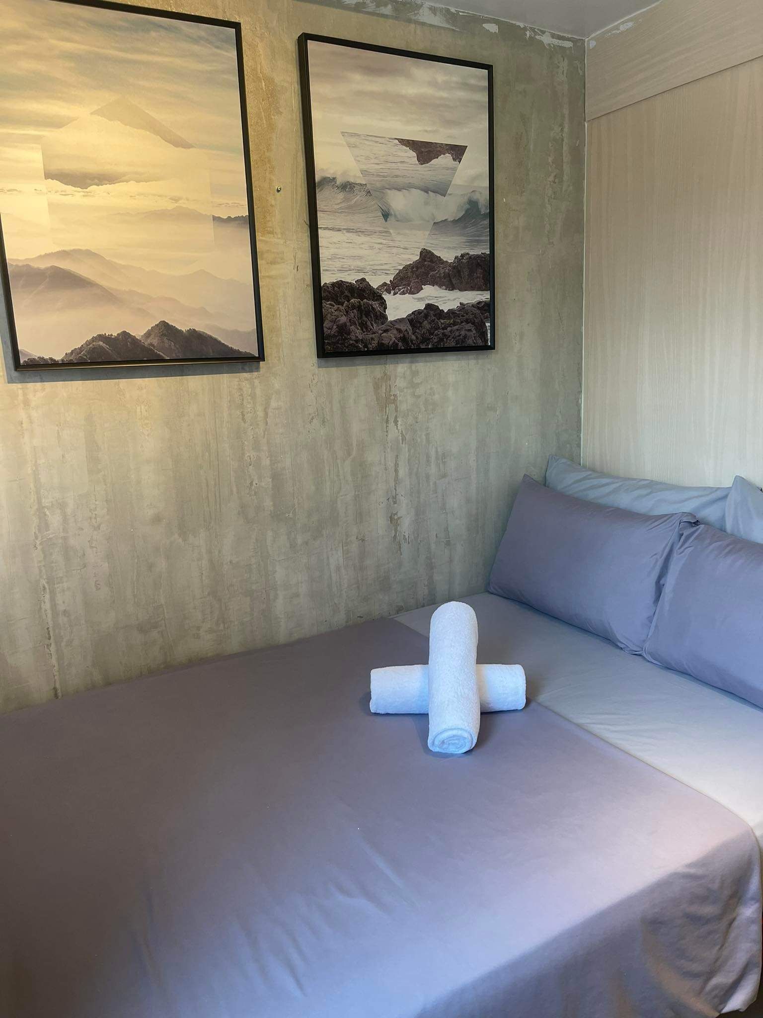 A hotel room with a bed, two framed pictures on the wall, and a rolled towel on the bed.