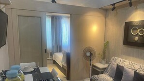 A modern hotel room with a bed, a sofa, a dining table with chairs, a standing fan, and a television mounted on the wall.