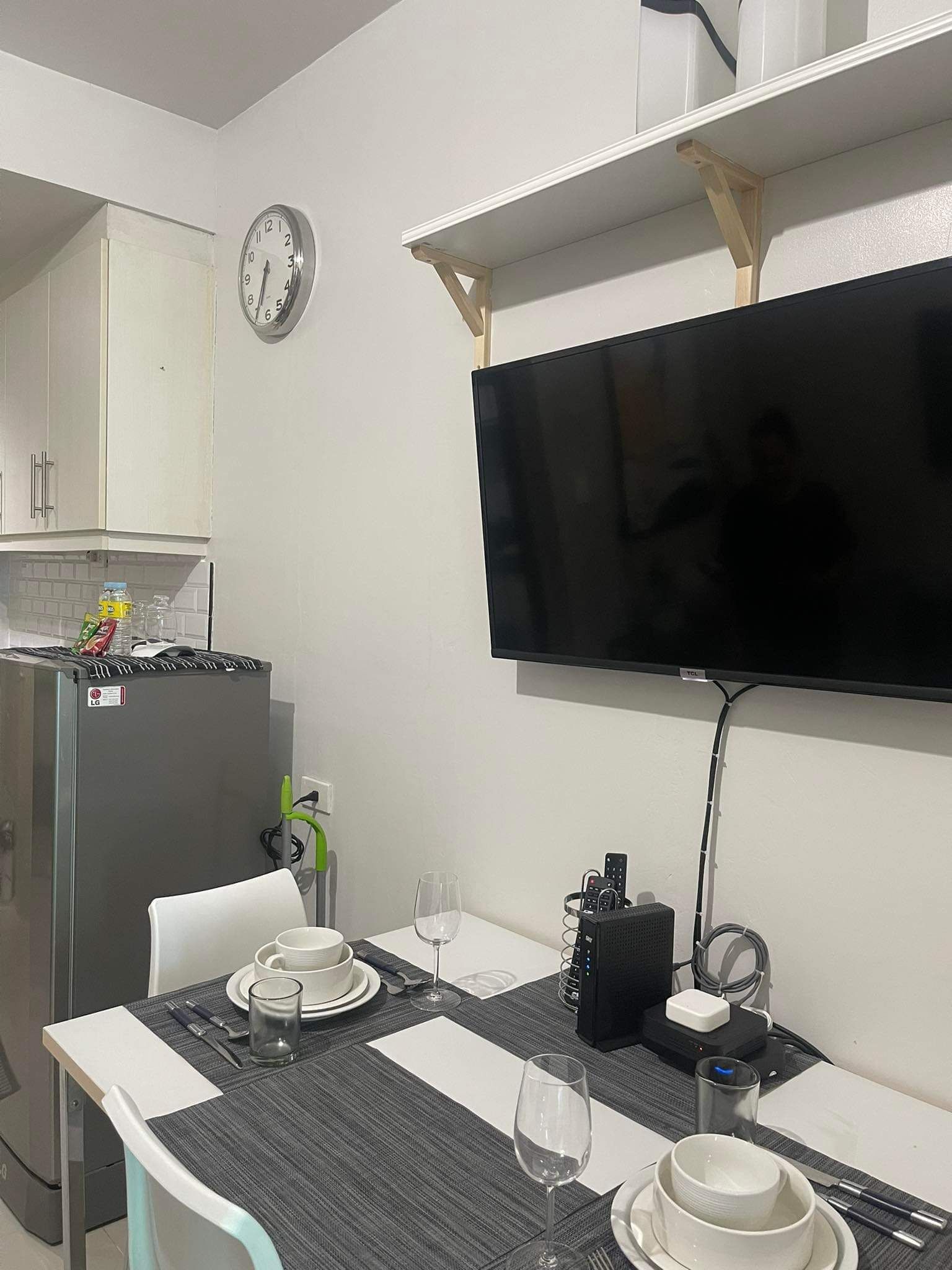 Et lille, moderne køkken med et køleskab, en mikrobølgeovn og et ur på væggen. Der er et spisebord med et TV ophængt over.