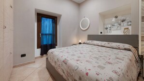 Quarto com cama, janela com cortinas, mesa de cabeceira, espelho fixado na parede e uma pequena estante com objetos decorativos.