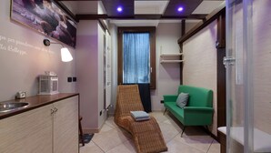 Quarto com cozinha compacta, cadeira de vime, sofá verde e janela com cortinas.
