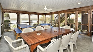 Une terrasse couverte avec une table en bois et des chaises blanches, un ventilateur de plafond et une vue sur l’extérieur.