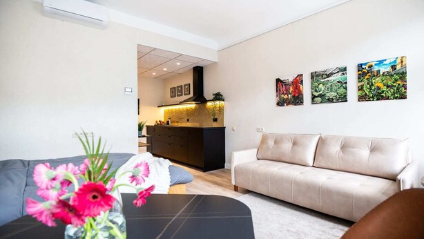 Un salon moderne avec un canapé, une table basse ornée d’un vase de fleurs et un coin cuisine visible à travers une porte ouverte.
