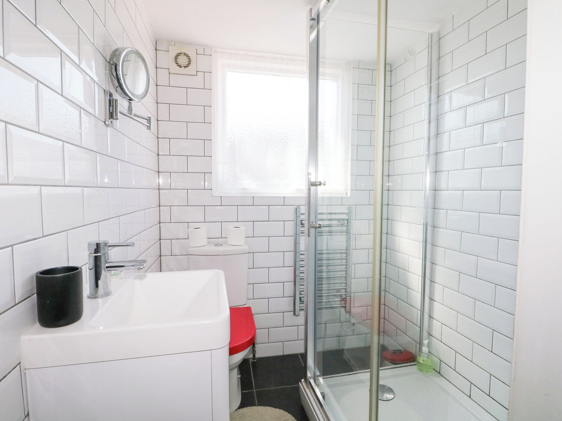Une salle de bain avec des murs recouverts de carreaux blancs, un lavabo blanc, des toilettes rouges, une cabine de douche en verre et une fenêtre laissant entrer la lumière naturelle.