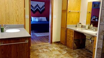 A bathroom with wooden cabinets, a sink, and a mirror. There is a door leading to a bedroom with a bed and a hanging decoration.