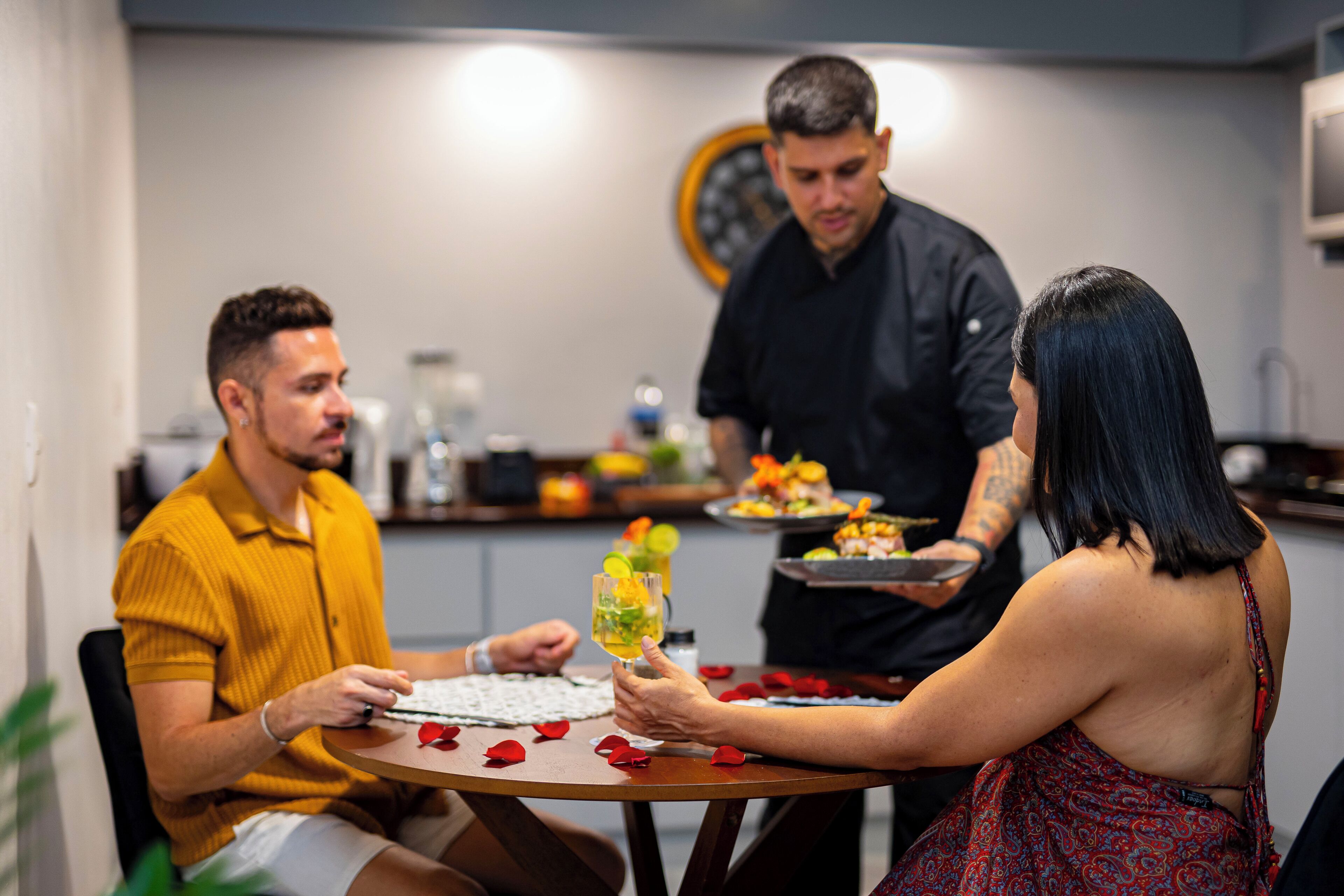 A dining table with a person holding a drink, another person seated, and a third person standing with a tray.