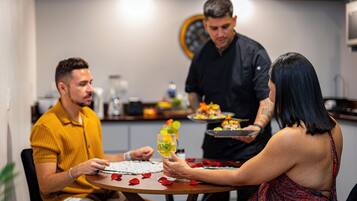 A dining table with a person holding a drink, another person seated, and a third person standing with a tray.