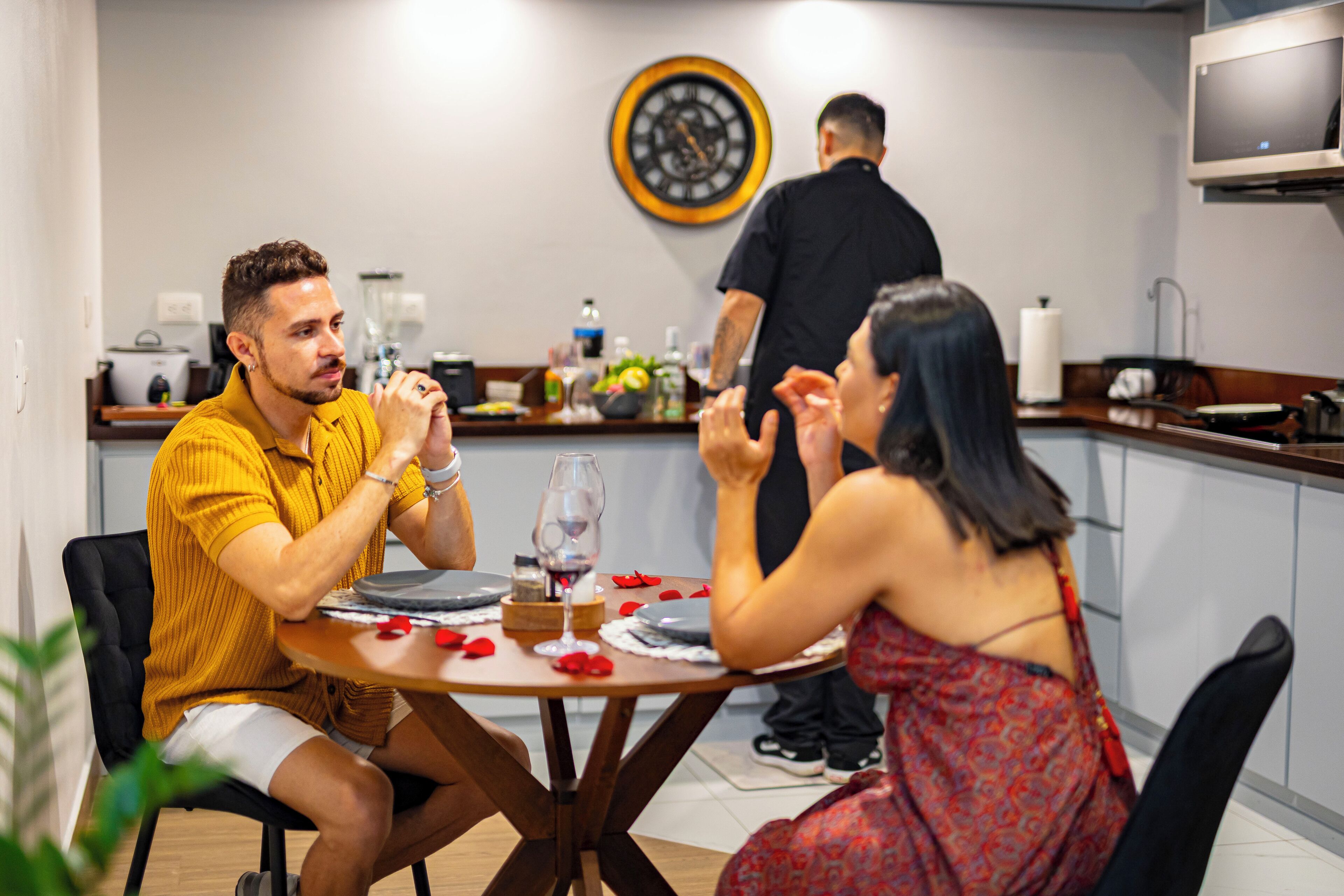 Un comedor con una mesa redonda, sillas, un reloj en la pared y una cocina al fondo.