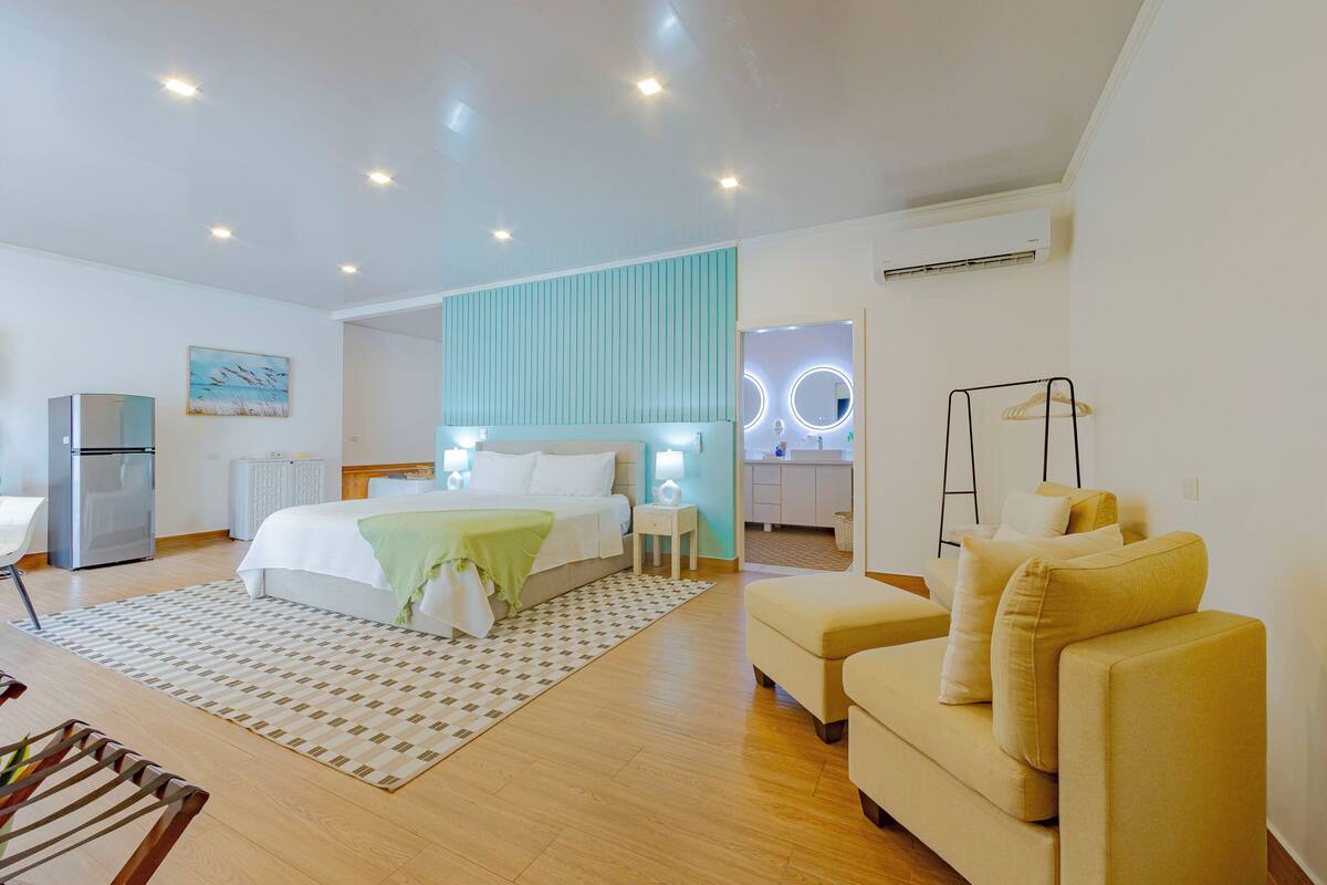A modern hotel room with a bed, bedside table, lamp, sofa, and a small kitchenette.