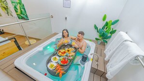 Una pareja en un jacuzzi, con una mesa de comida y bebidas, rodeada de plantas y un toallero blanco.