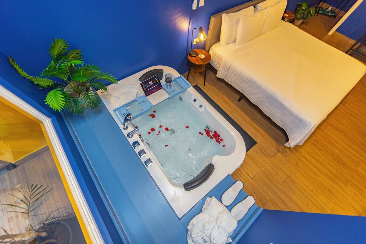 A room with a bathtub, a bed, a small table with a lamp, and a potted plant.