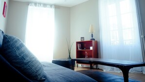 A living room with a sofa, a table, a red bookshelf, and a window with sheer curtains. - Chez Armande 2-star tourist apartment 48 m2 (Châtel-Guyon)