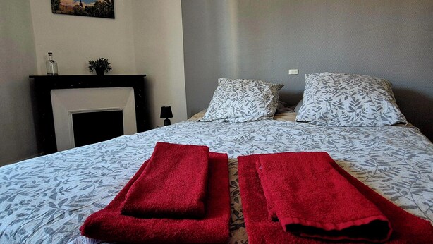 A hotel room with a bed, two red towels, a nightstand, a lamp, a painting on the wall, and a window with curtains. - Chez Armande 2-star tourist apartment 48 m2 (Châtel-Guyon)