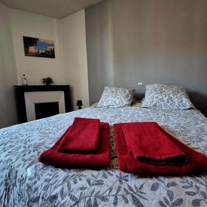 A hotel room with a bed, two red towels, a nightstand, a lamp, a painting on the wall, and a window with curtains. - Chez Armande 2-star tourist apartment 48 m2 (Châtel-Guyon)