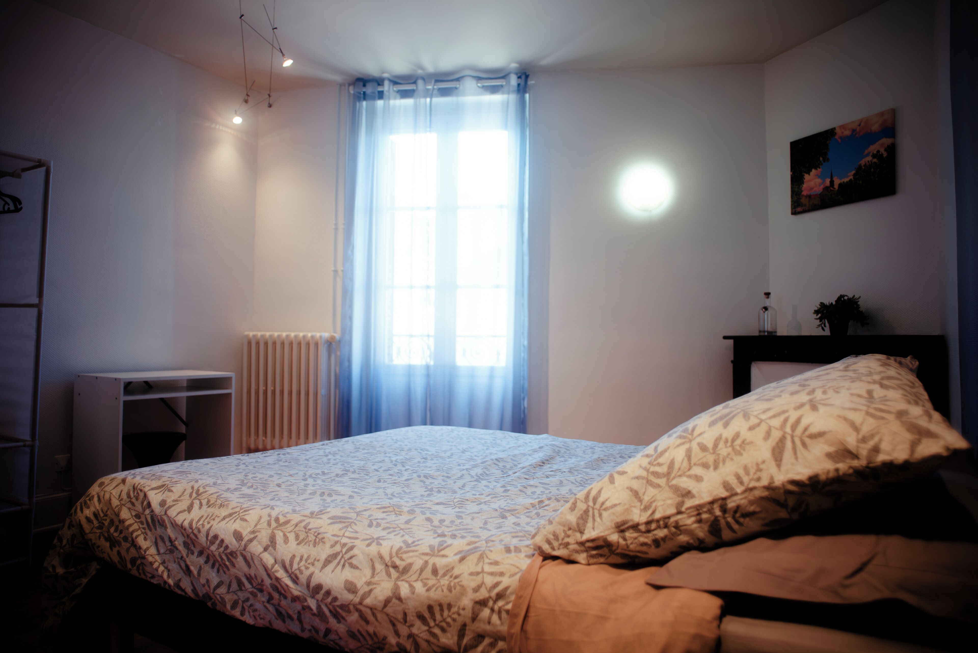 Un dormitori amb un llit, un radiador, una finestra amb cortines, un escriptori i un quadre penjat a la paret.