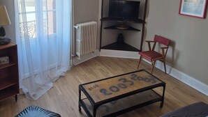 A living room with a wooden floor, a glass coffee table, a black ottoman, a red chair, a TV on a stand, and a radiator. - Chez Armande 2-star tourist apartment 48 m2 (Châtel-Guyon)