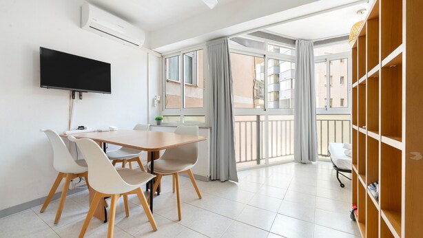 Una sala de estar moderna con una televisión de pantalla plana, una mesa de comedor con sillas, una estantería y un balcón con cortinas.