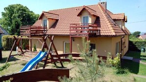 A two-story house with a red-tiled roof, a wooden swing set, and a slide in the yard. - Holiday Apartment for 4 People, Lubin (Międzyzdroje)