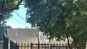 En lukket indgang med et metalhegn, et træ i forgrunden og en bygning med en hvid facade i baggrunden.