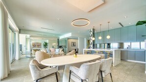 A modern kitchen with a large island, pendant lighting, and a dining area with a round table and chairs. - Laguna Del Mar - Prime Beachfront Luxury (Grand Cayman)