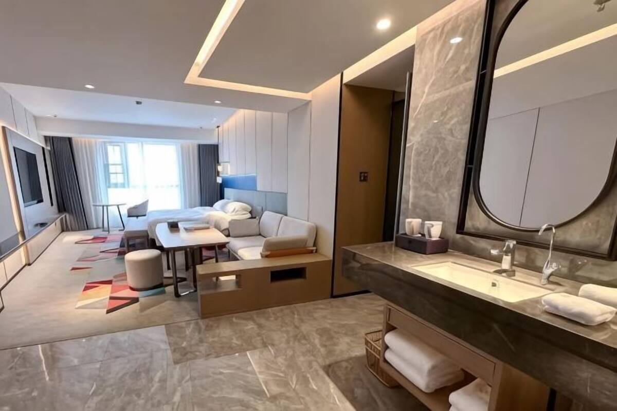 Een moderne hotelkamer met een groot bed, een zithoek met een bank en een salontafel, een badkamer met een grote spiegel en wastafel, en een uitzicht op de stad.