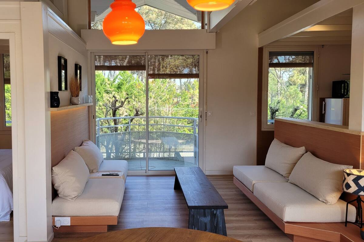 Una habitación moderna con cama, sofá y mesa de comedor, ventanas amplias y un balcón con vistas a los árboles.