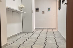 Un couloir avec un sol carrelé, une porte blanche et une étagère sur laquelle est posé un vase de fleurs.