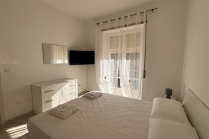 Ein Schlafzimmer mit einem Bett, einem Schminktisch, einem an der Wand montierten Fernseher und einem Fenster mit durchsichtigen Vorhängen.