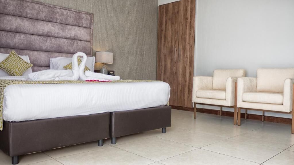 A modern hotel room with a large bed, two armchairs, a wooden wardrobe, and a lamp.
