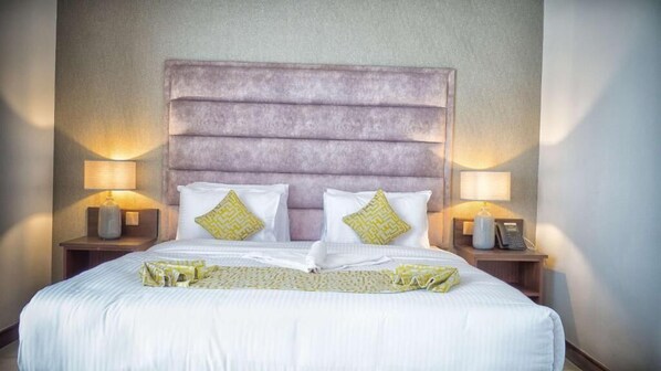 A neatly made bed with a headboard, two bedside lamps, and a bedside table with a phone and a vase. - FAIRWAYS HOTEL KISUMU (Kisumu)
