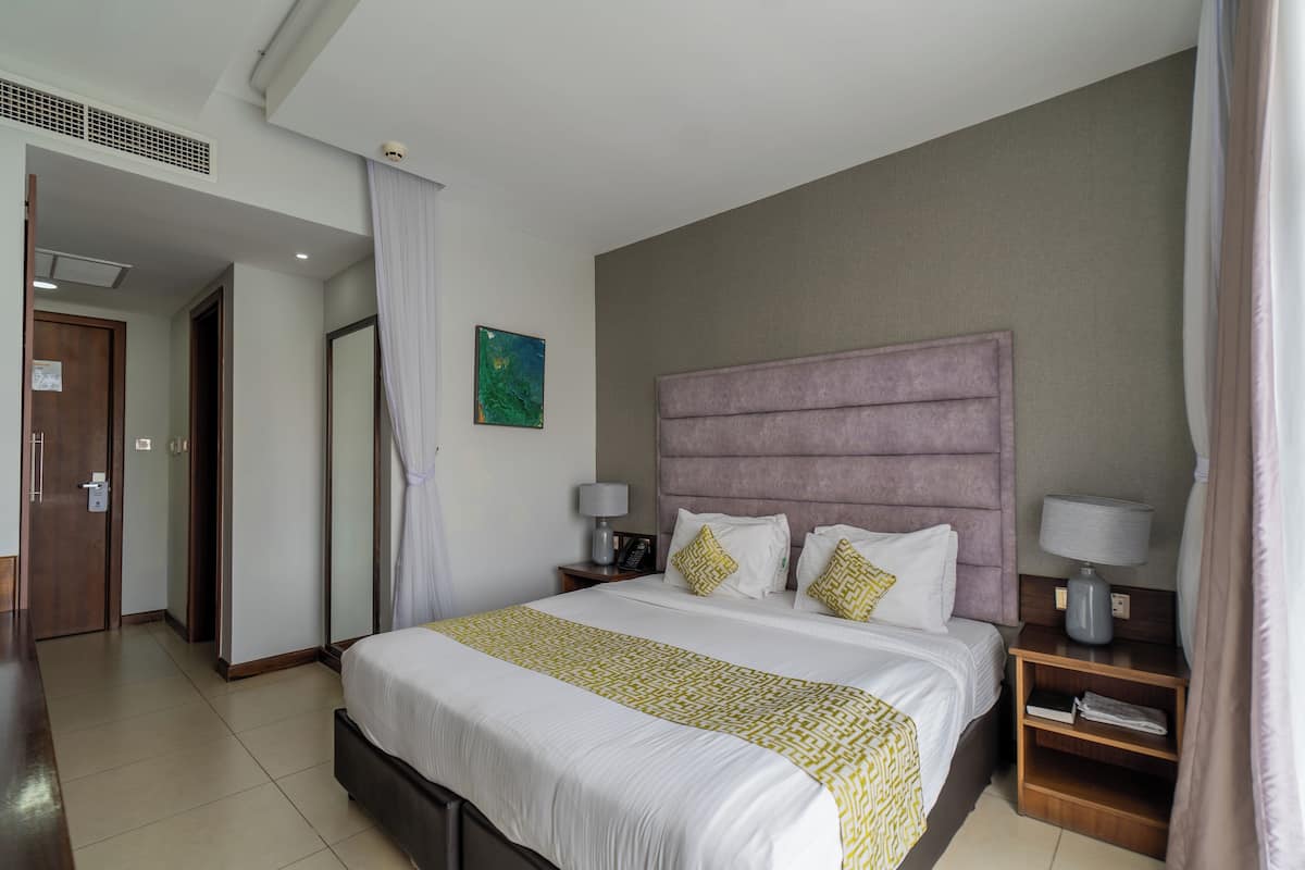 Une chambre d’hôtel avec un grand lit, deux tables de chevet avec des lampes, une tête de lit, un tableau accroché au mur et une porte donnant sur une autre pièce.