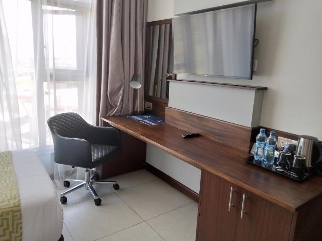 Habitación de hotel con escritorio de madera, una silla de oficina negra, un televisor montado sobre el escritorio y una ventana con cortinas.