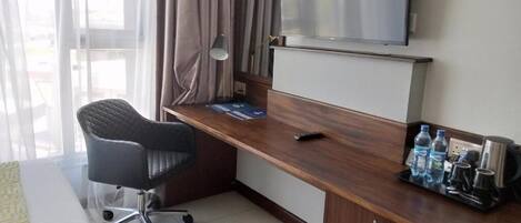A hotel room with a wooden desk, a black office chair, a TV mounted above the desk, and a window with curtains.