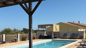 A pool area with lounge chairs, a pergola, and a building with a tiled roof.