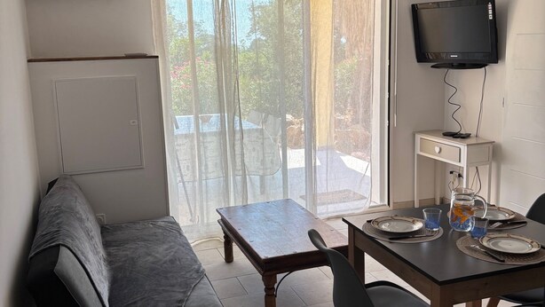 A compact living space with a sofa, a dining table set for two, a TV mounted on the wall, and a window with sheer curtains.