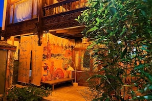 A traditional wooden building with a mural, a bench, and lush greenery. - Wu Suo · Li Ziyuan (Lijiang Old Town Mufu Hotel) (Lijiang)