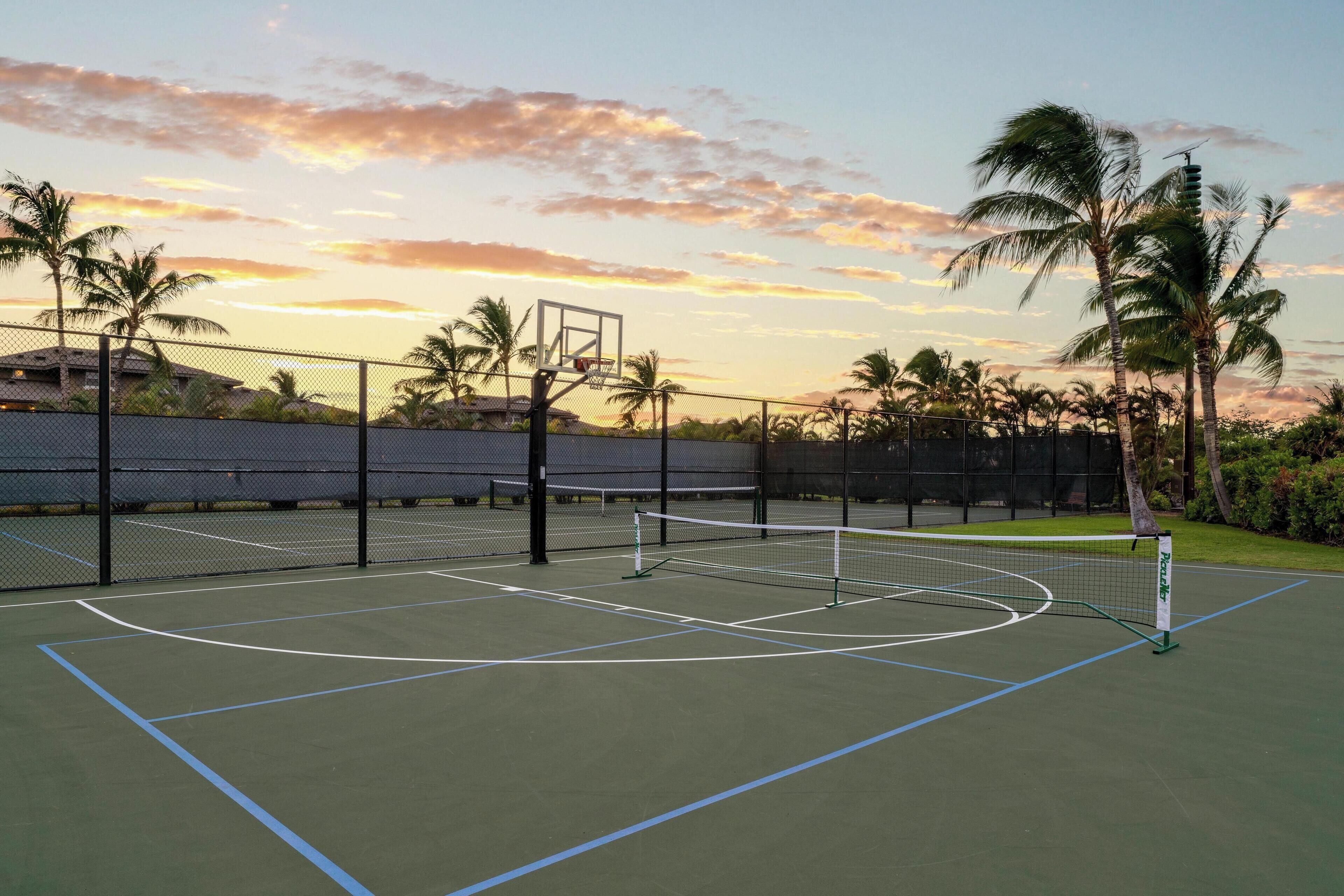 Una cancha de tenis exterior con red, rodeada por una valla, palmeras y un atardecer de fondo.