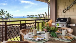 A covered outdoor dining area with a view of the ocean and palm trees, a grill, and a table set for two with a vase of flowers. - Ocean Views!  8A Halii Kai (Waikoloa Beach Resort)