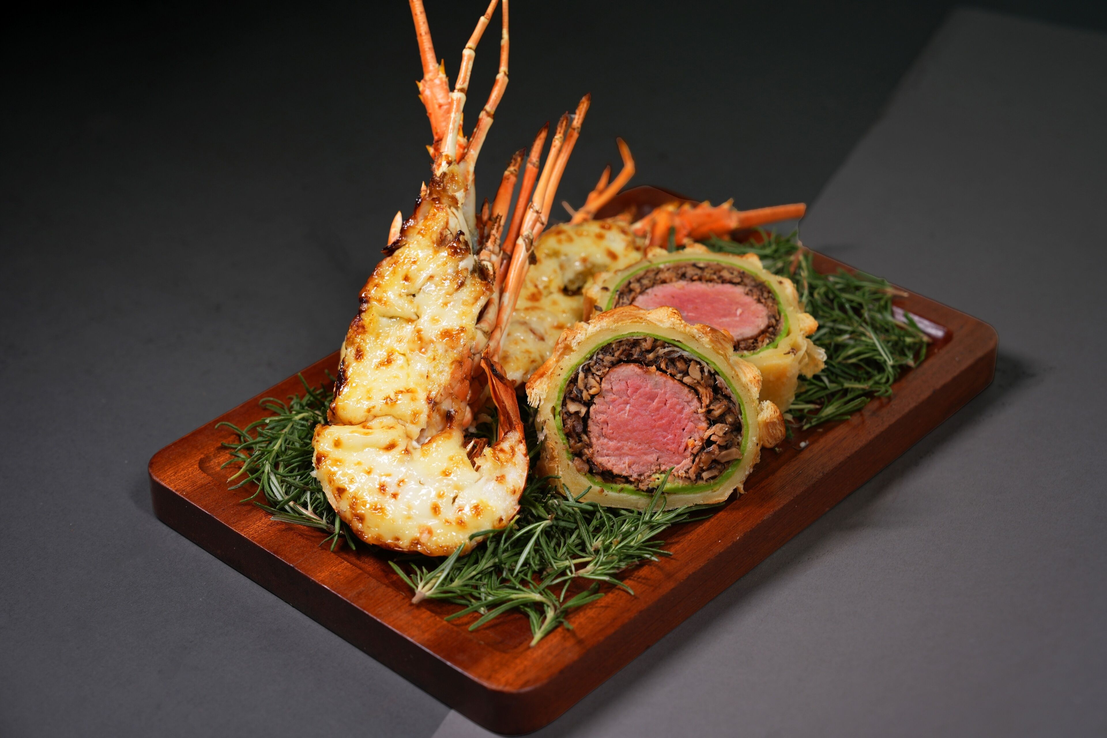 A lobster tail, a rolled meat dish, and a stuffed vegetable on a wooden board.