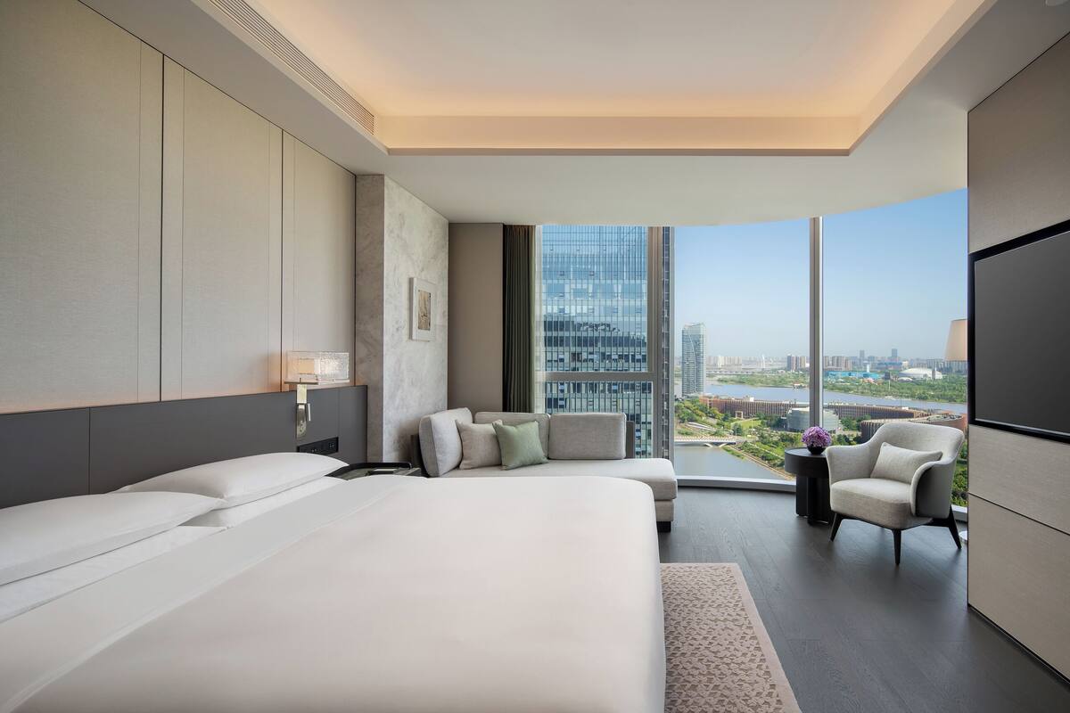 A modern hotel room with a large bed, a sofa, a chair, and a TV. The room has a large window with a city view.