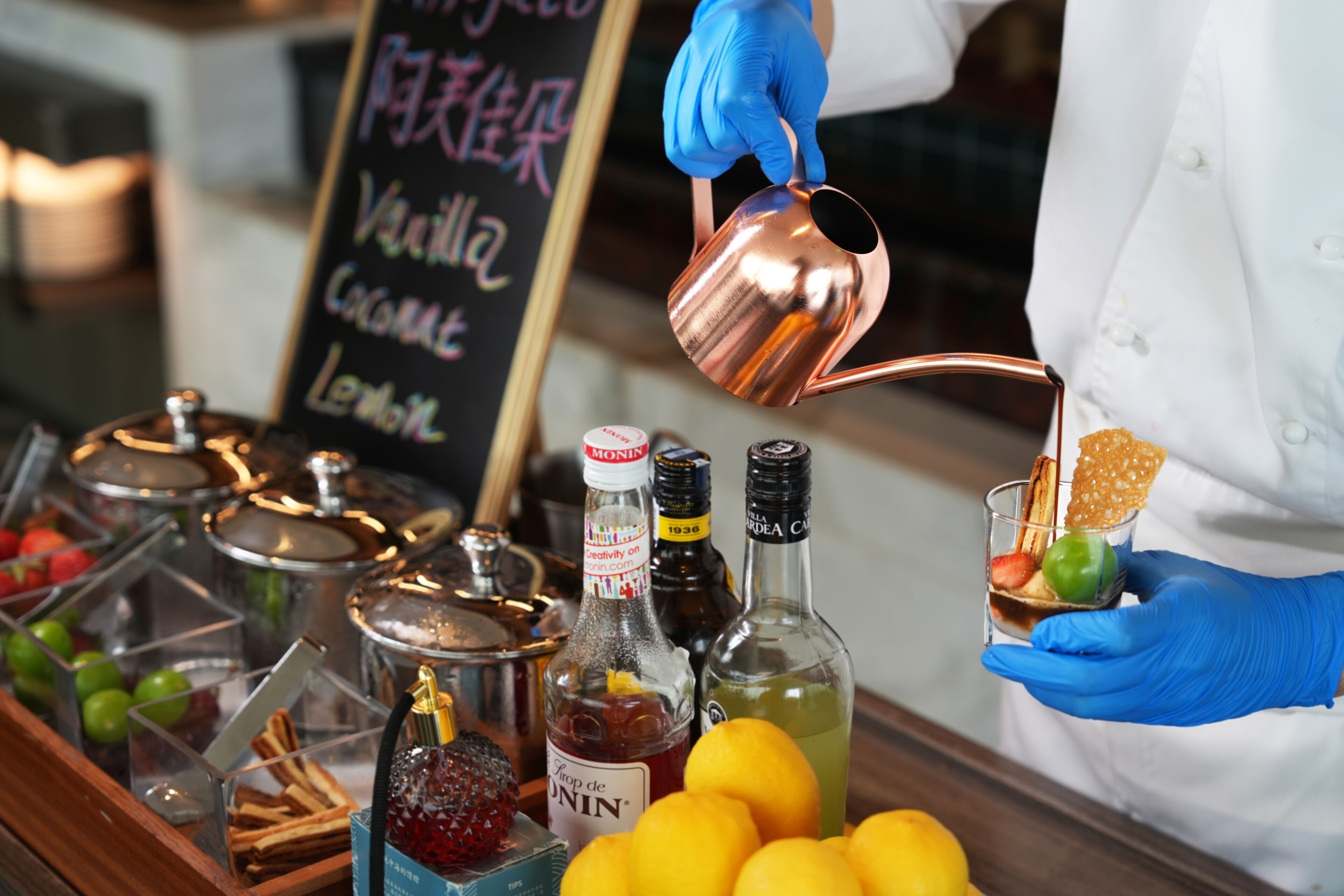 A person in a white coat and blue gloves pouring a liquid from a copper jug into a glass containing a green fruit and a wafer.