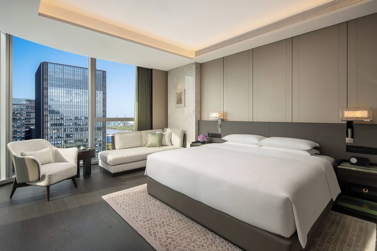 A modern hotel room with a large bed, a sofa, a chair, and a view of a cityscape through large windows.