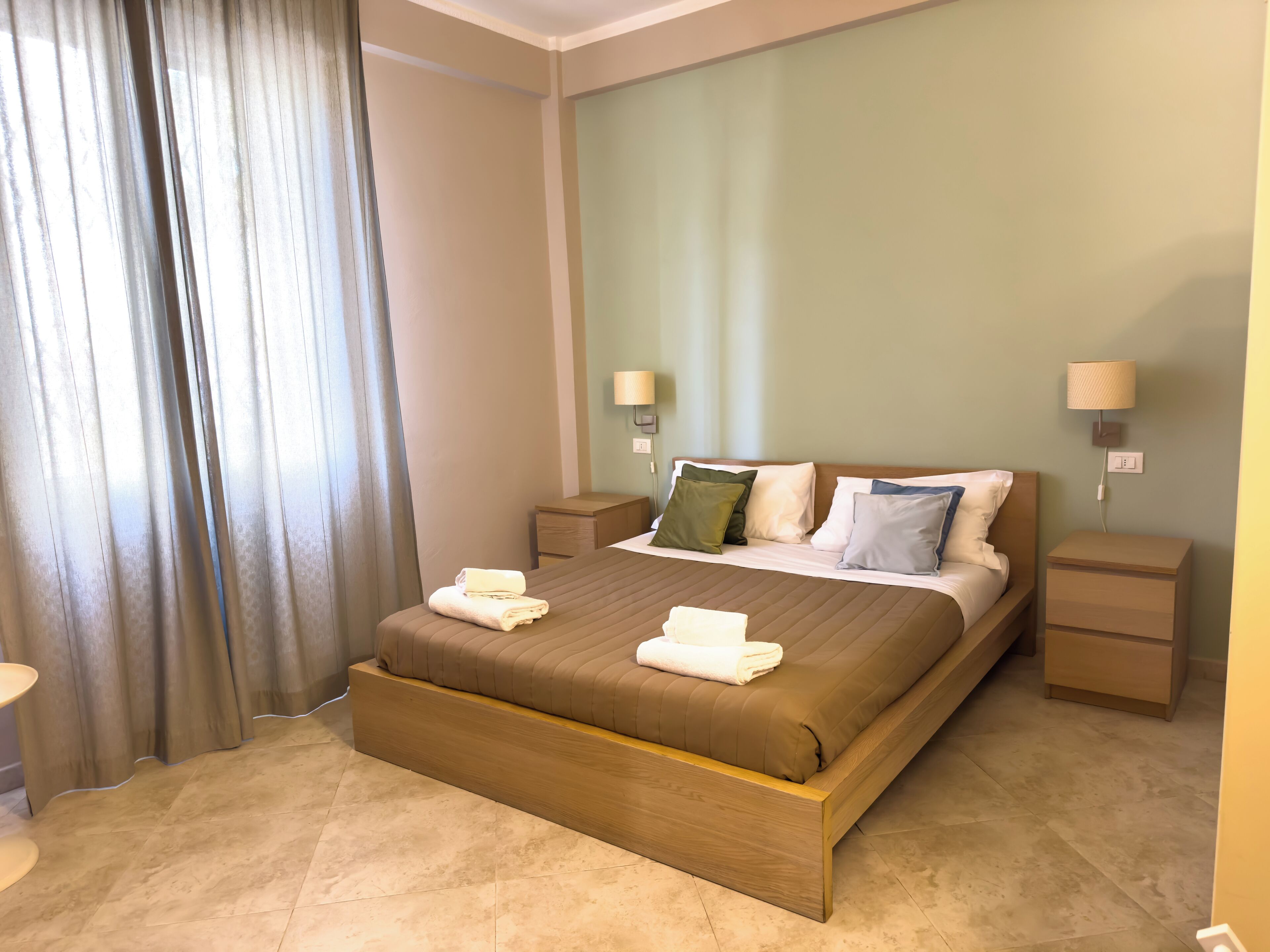 A hotel room with a large bed, two bedside tables, a nightstand, a lamp, and a window with curtains.